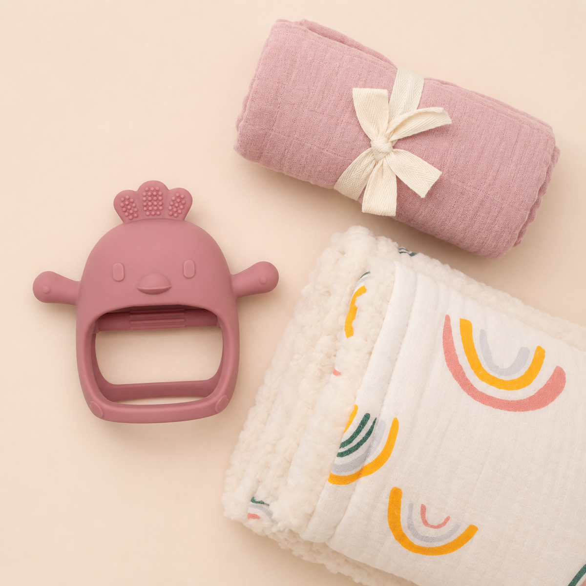 Product flat lay of baby pink bamboo swaddle, rainbow print sherpa muslin blanket, deep pink baby teether