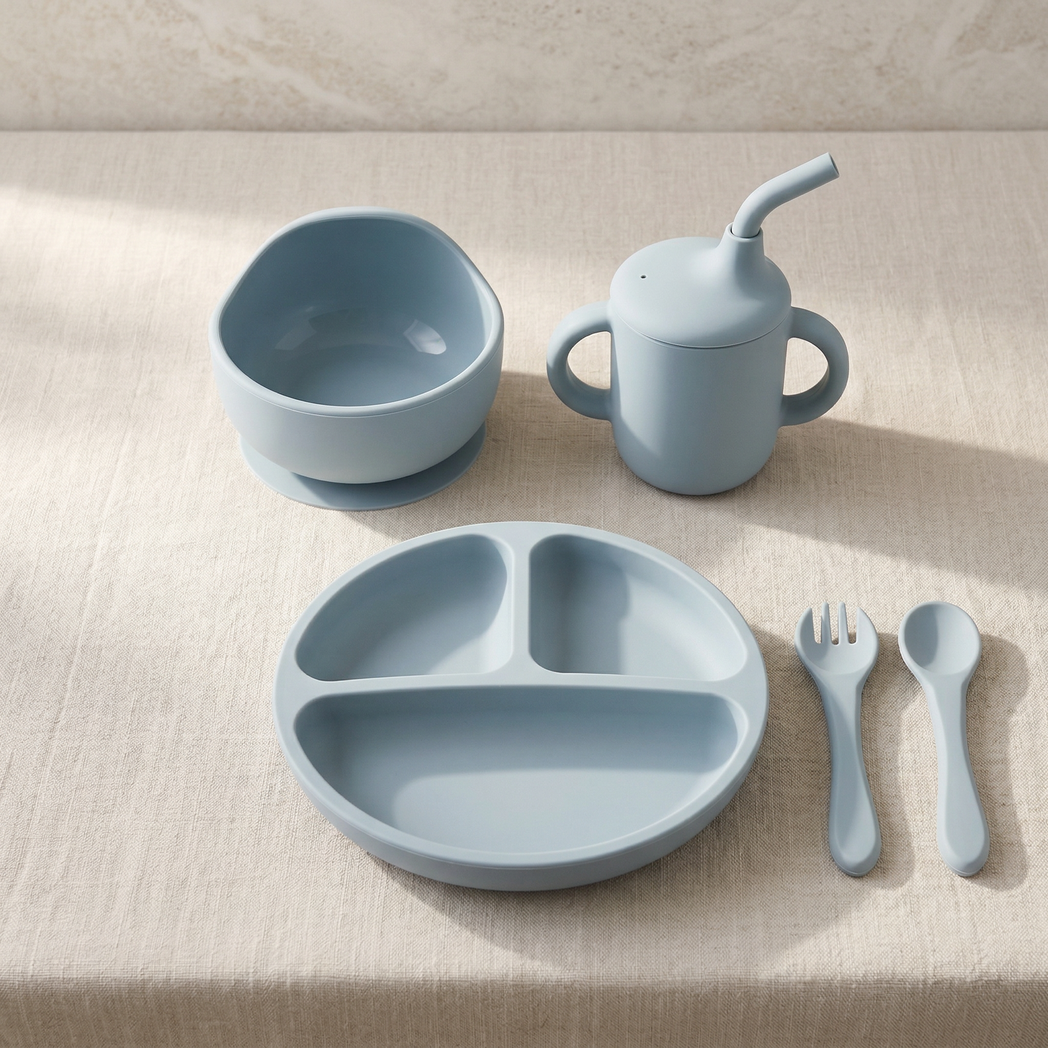 Set of blue children's dining utensils including a bowl, cup, plate, fork, spoon, and straw on a beige surface.
