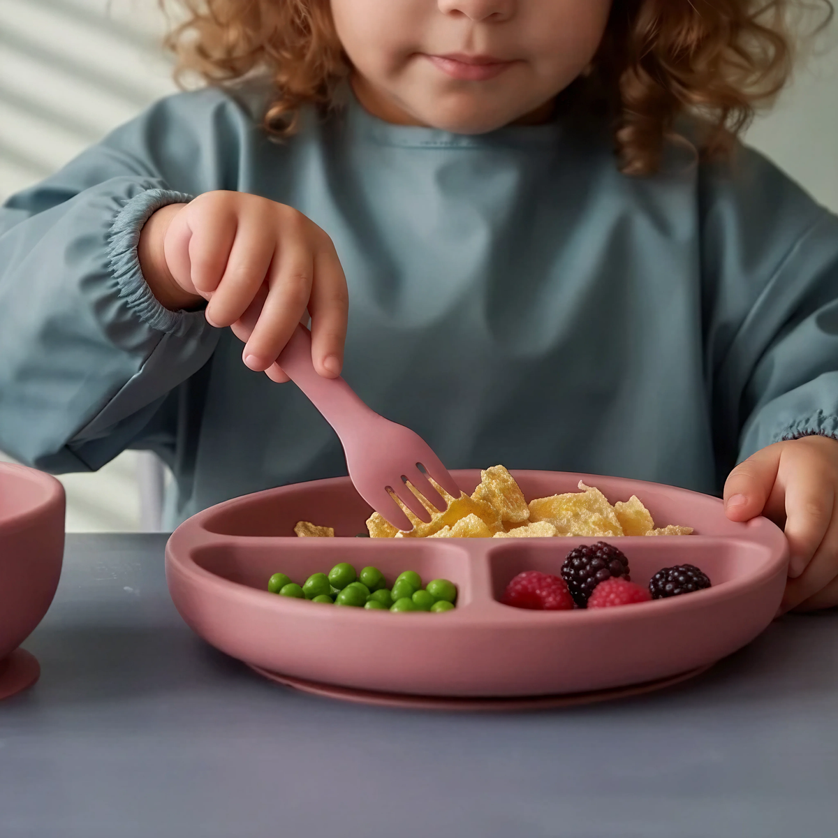 Toddler girl using 5 piece baby silicone feeing set in Rosewood (blush pink) with suction plate, sippy cup, suction bowl, fork and spoon