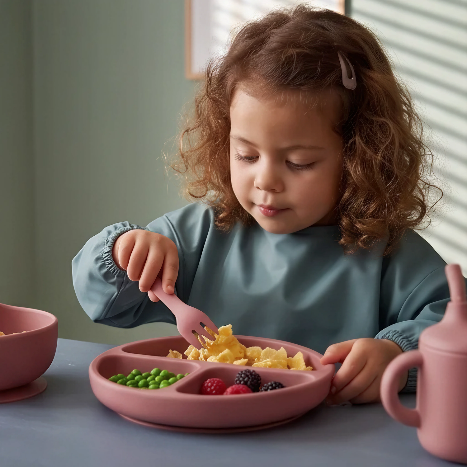 Toddler girl using 5 piece baby silicone feeing set in Rosewood (blush pink) with suction plate, sippy cup, suction bowl, fork and spoon