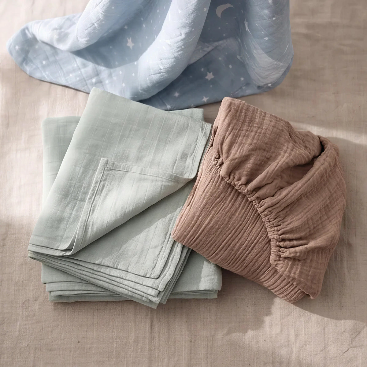 Sea Mist muslin swaddle and Cocoa fitted cot sheet flat lay with blue lunar star print - Honey & Sage