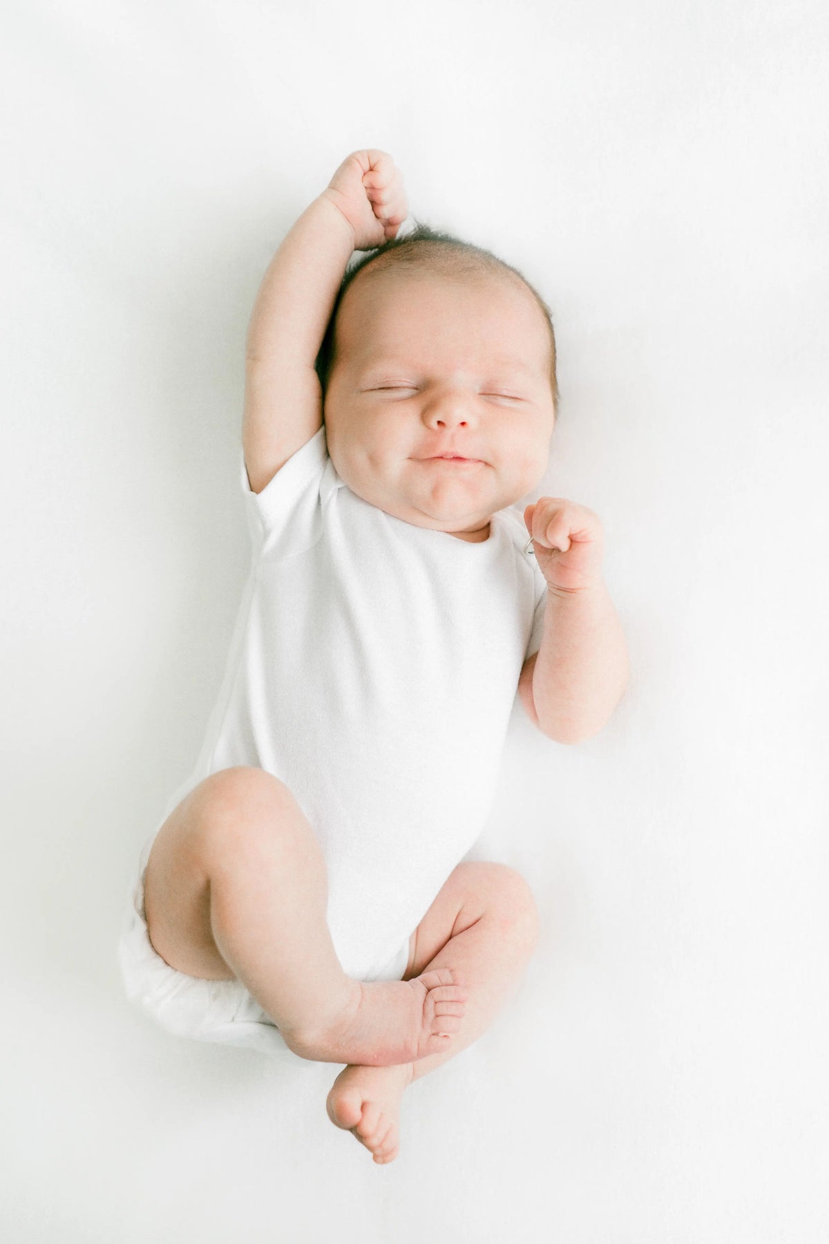 Newborn baby asleep in a white bodysuit with arms stretched overhead